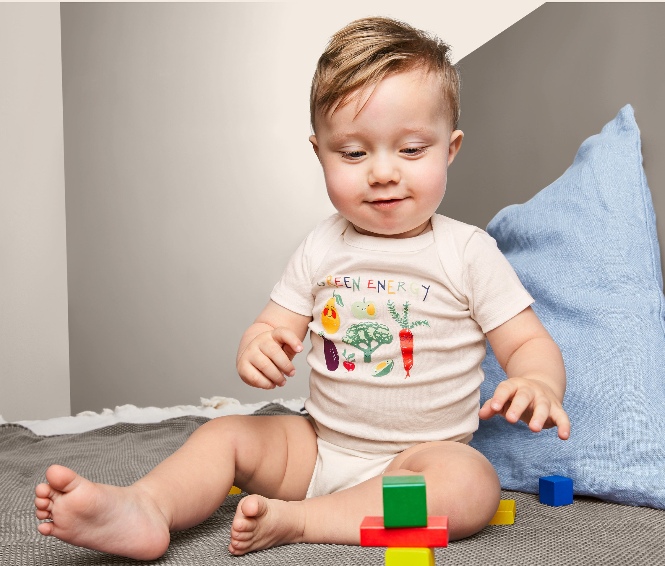 Baby sitzt auf einer Decke und spielt mit bunten Würfeln. Es trägt einen 3 Kurzarmbody mit Gemüseaufdruck.