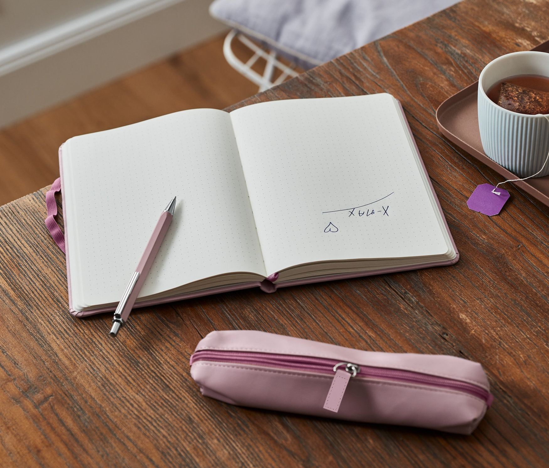 Offenes Notizbuch mit Stift und Etui liegt auf einem Holztisch neben einer Tasse Tee und Keksen.