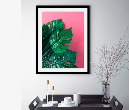 Ein gerahmtes Bild »Monstera Pertusum«, 30 x 40 cm, hängt über einem schwarzen Tisch mit einer Vase und Kerzen.