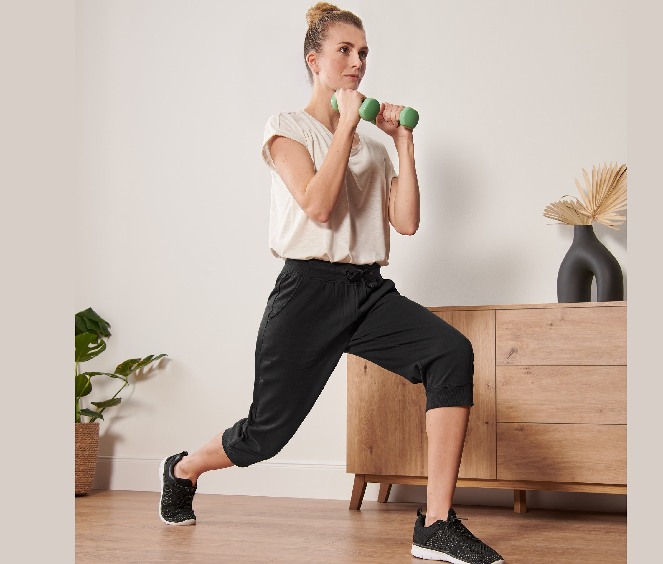 Frau macht Ausfallschritte mit grünen Hanteln und trägt eine schwarze 3/4-Sporthose.