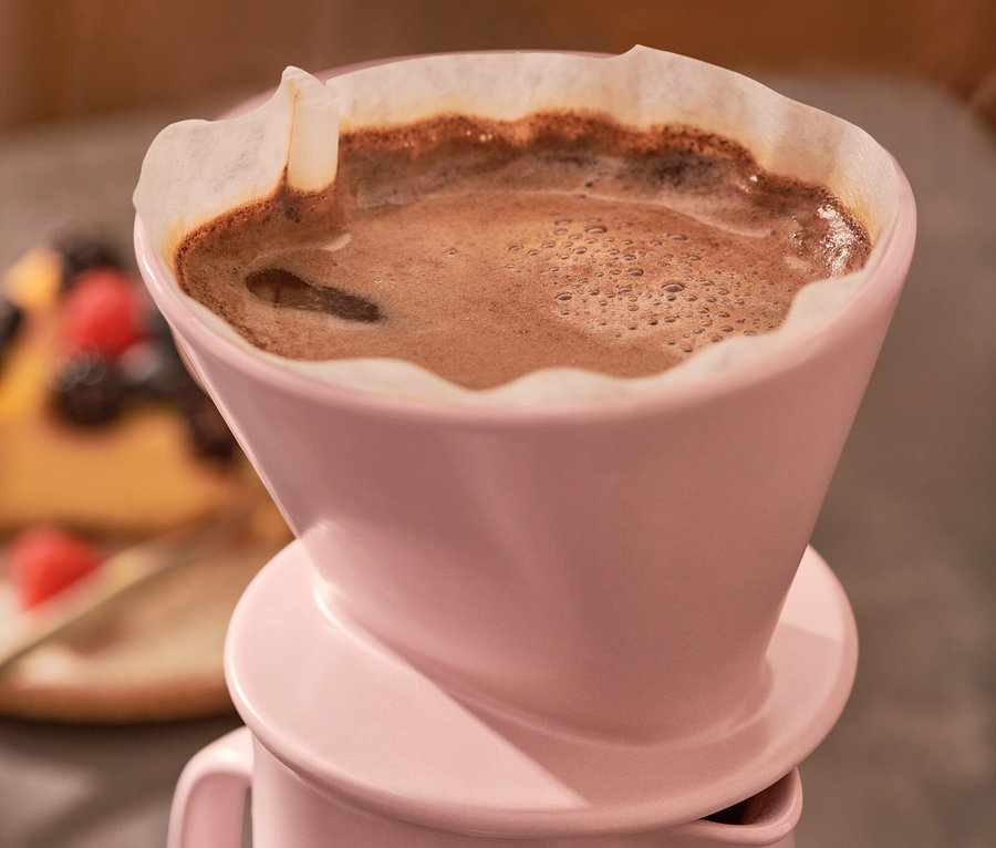 Nahaufnahme einer rosa Kaffeekanne mit Filter, aus der Dampf aufsteigt, mit einem Stück Kuchen im Hintergrund.