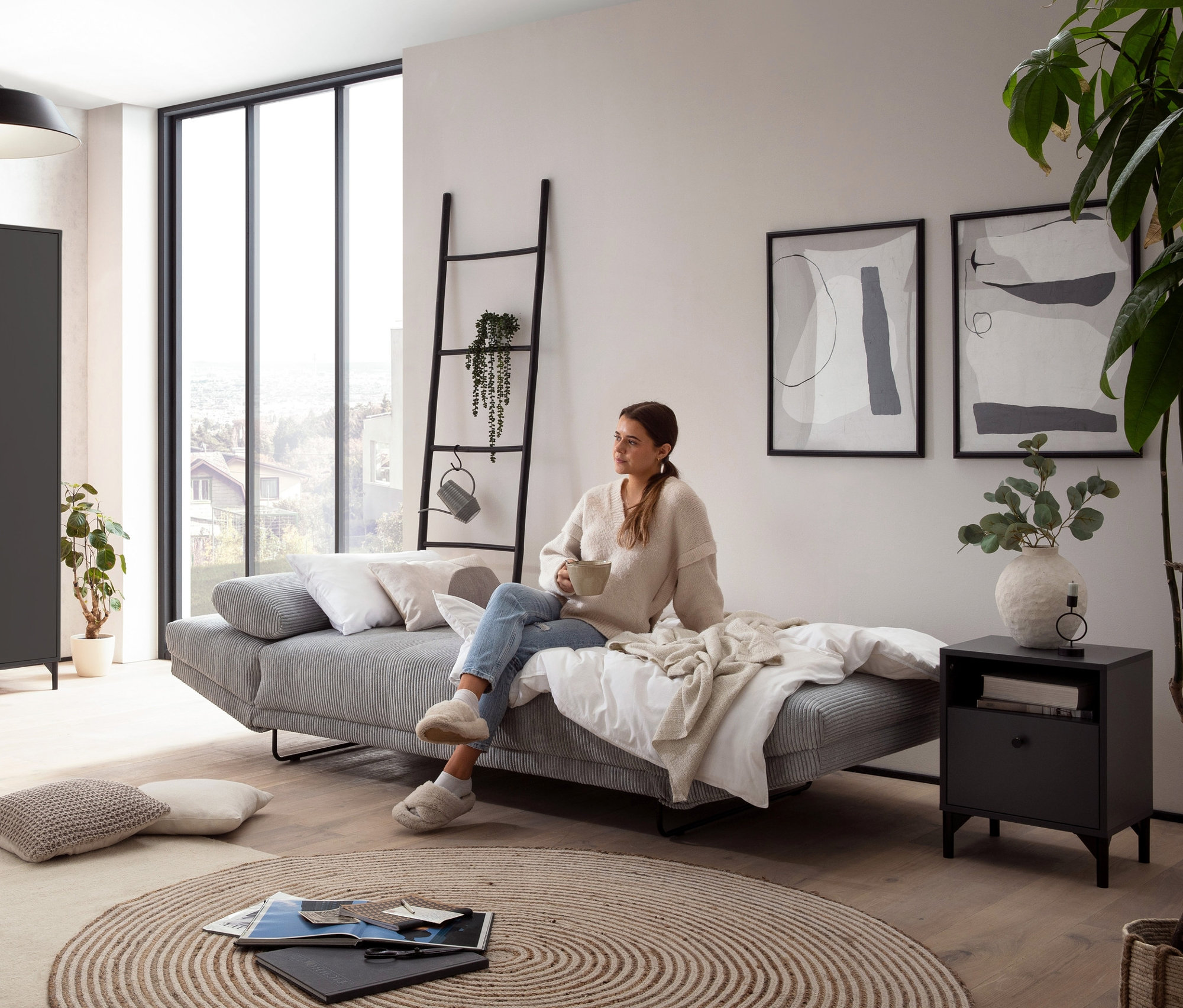 Eine Frau sitzt auf einem Sofa im Wohnzimmer. Im Hintergrund sind eine anthrazitfarbene Nachtkonsole, ein Schrank und ein Regal „Smash“ zu sehen.