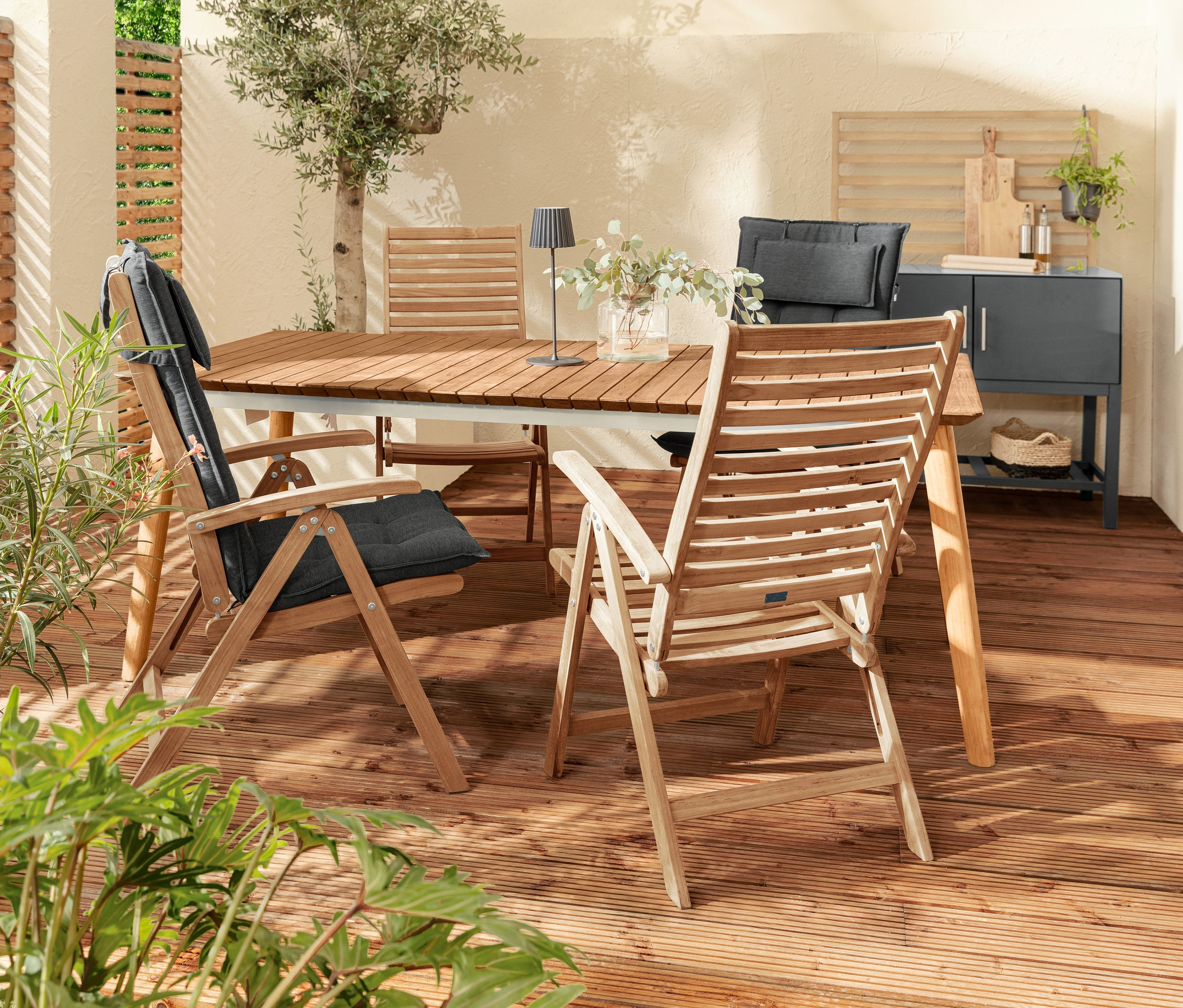 Außenterrasse mit Teak-Ausziehtisch, Stühlen mit anthrazitfarbenen Premium-Hochlehnerauflagen und schwarzer LED-Akku-Tischleuchte. Im Hintergrund Outdoor-Küchenmodul mit Lamellen-Wand.