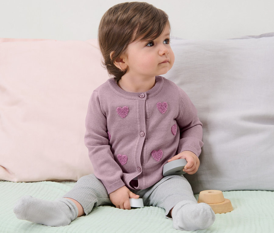 Kleinkind sitzt auf einem Bett und trägt eine Kinder-Strickjacke.