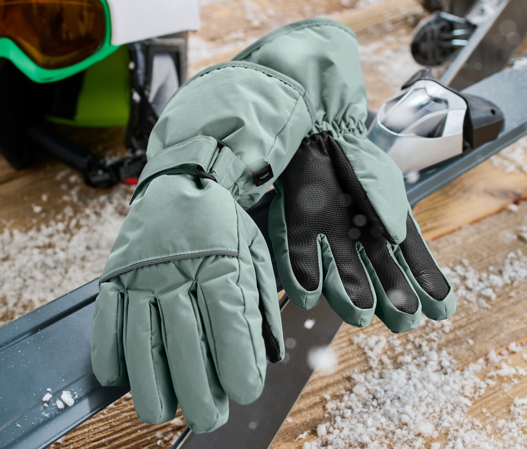 Grüne Kinder-Ski- und Winterhandschuhe liegen auf Skiern neben Helm und Brille.
