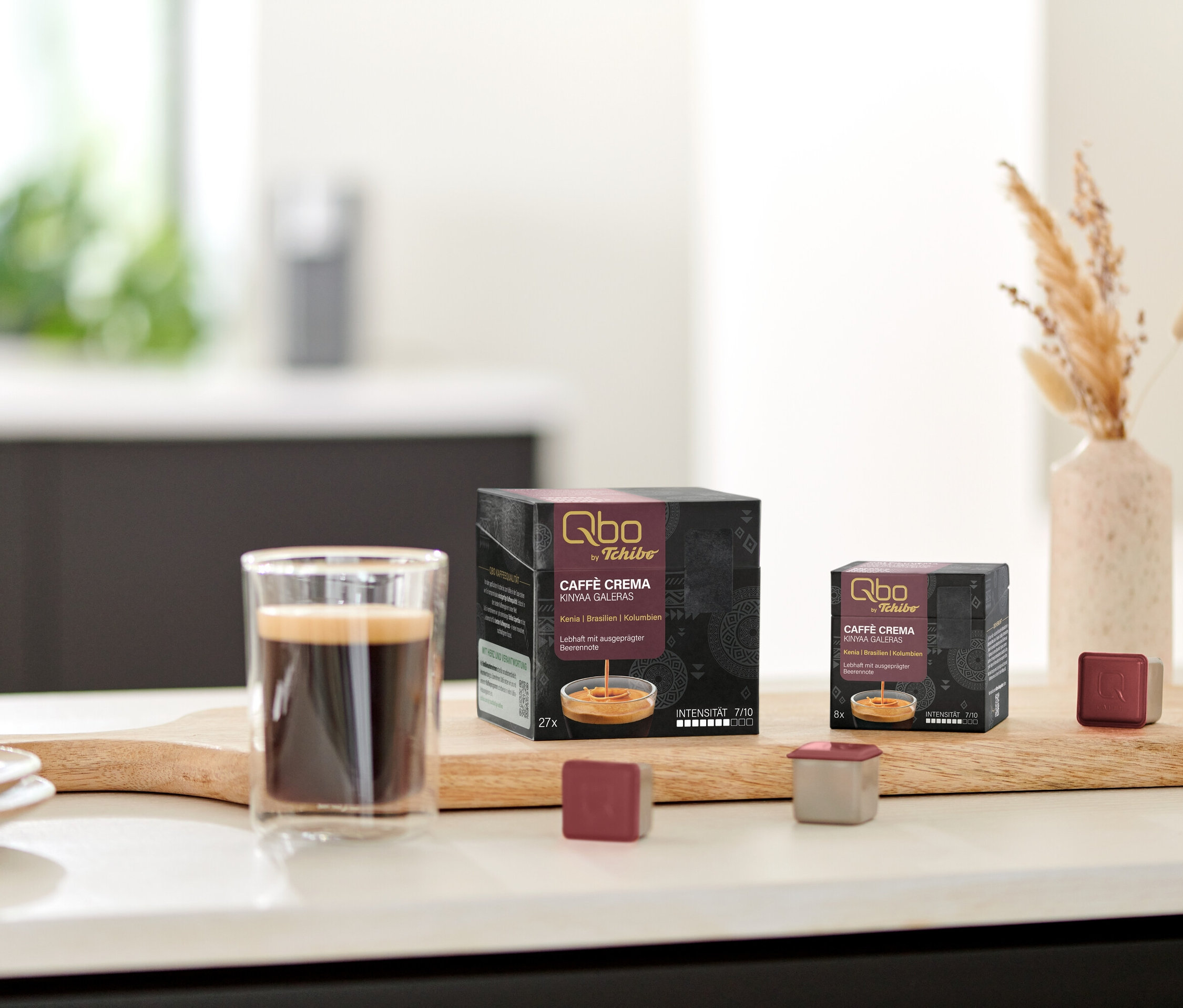 Ein Glas Kaffee steht neben Qbo Caffè KINYAA GALERAS Kapselpackungen auf einem Holzbrett.