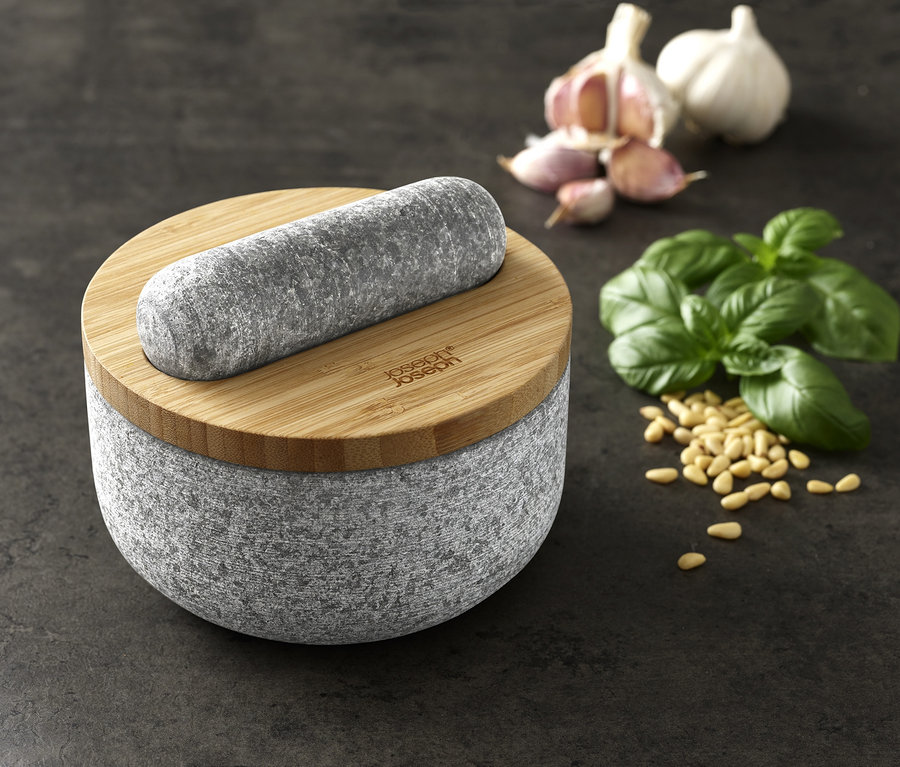Joseph Joseph Granit Mörser »Dash« mit Stößel auf Holzdeckel, Knoblauch, Basilikum und Pinienkerne auf dunklem Hintergrund.