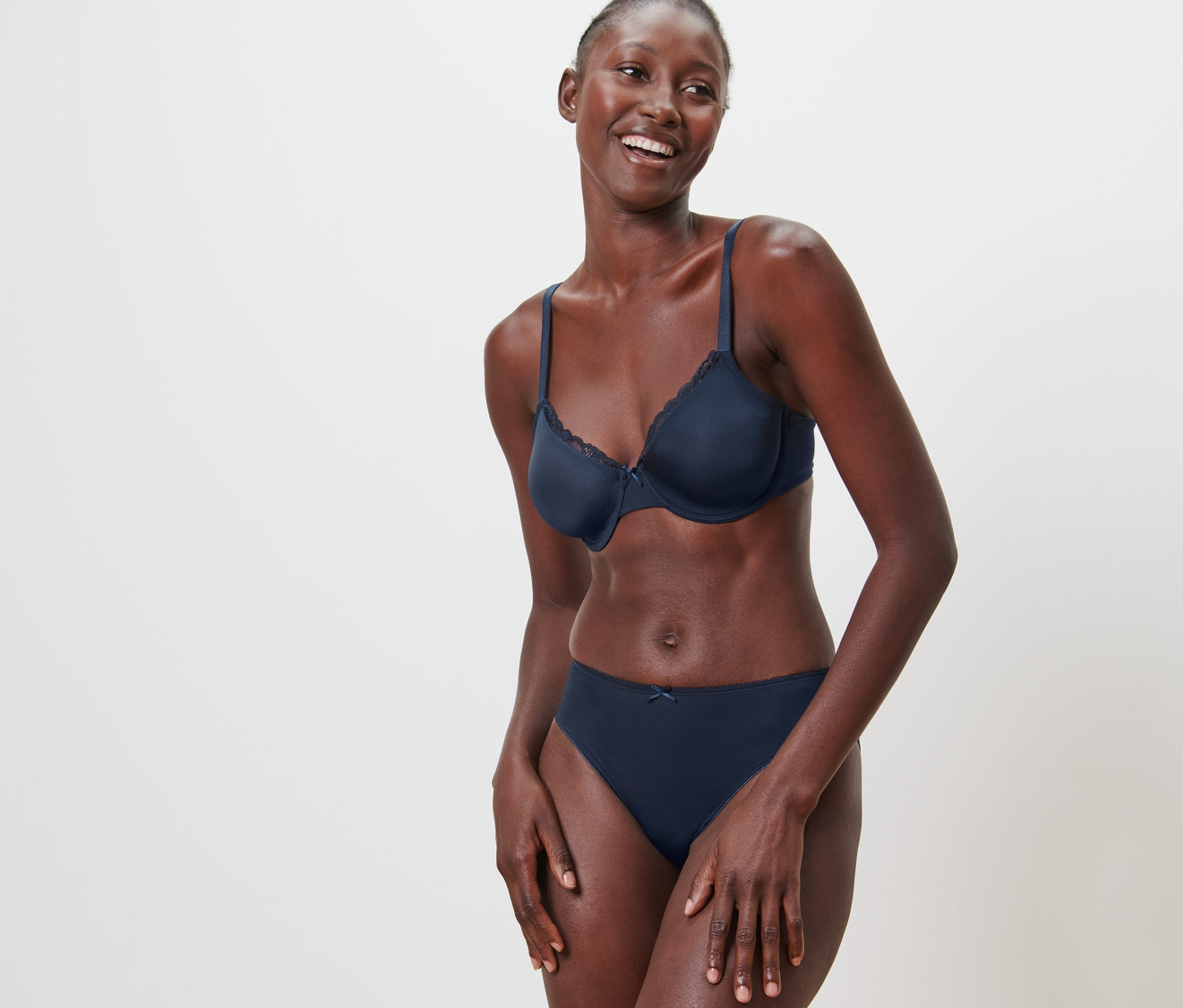 Lächelnde Frau posiert in Bügel-BH und Unterhose vor einem weißen Hintergrund.