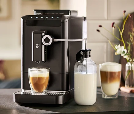 Ein »Esperto 2 Milk« Granit Black Kaffeevollautomat bereitet Kaffee in einem Glas zu. Daneben steht eine Milchkaraffe.