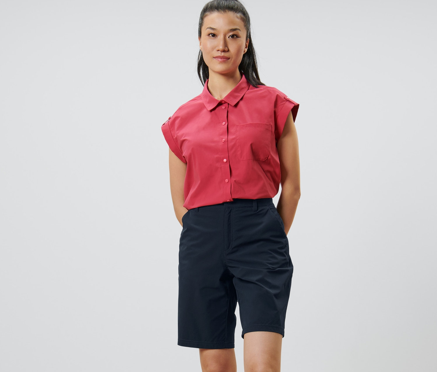 Eine Frau steht mit den Händen hinter dem Rücken. Sie trägt dunkelblaue Softshell-Funktionsshorts und ein rotes Hemd.