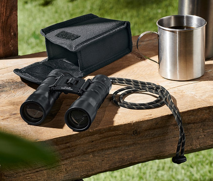 Ein »12x32«-Taschenfernglas liegt neben einem Etui, einer Thermoskanne und einer Tasse auf einer Holzbank.