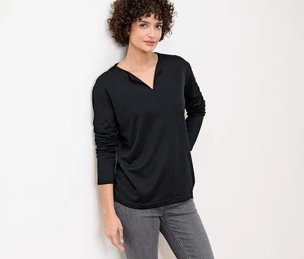 Frau lehnt an Wand, trägt ein schwarzes Blusenshirt.