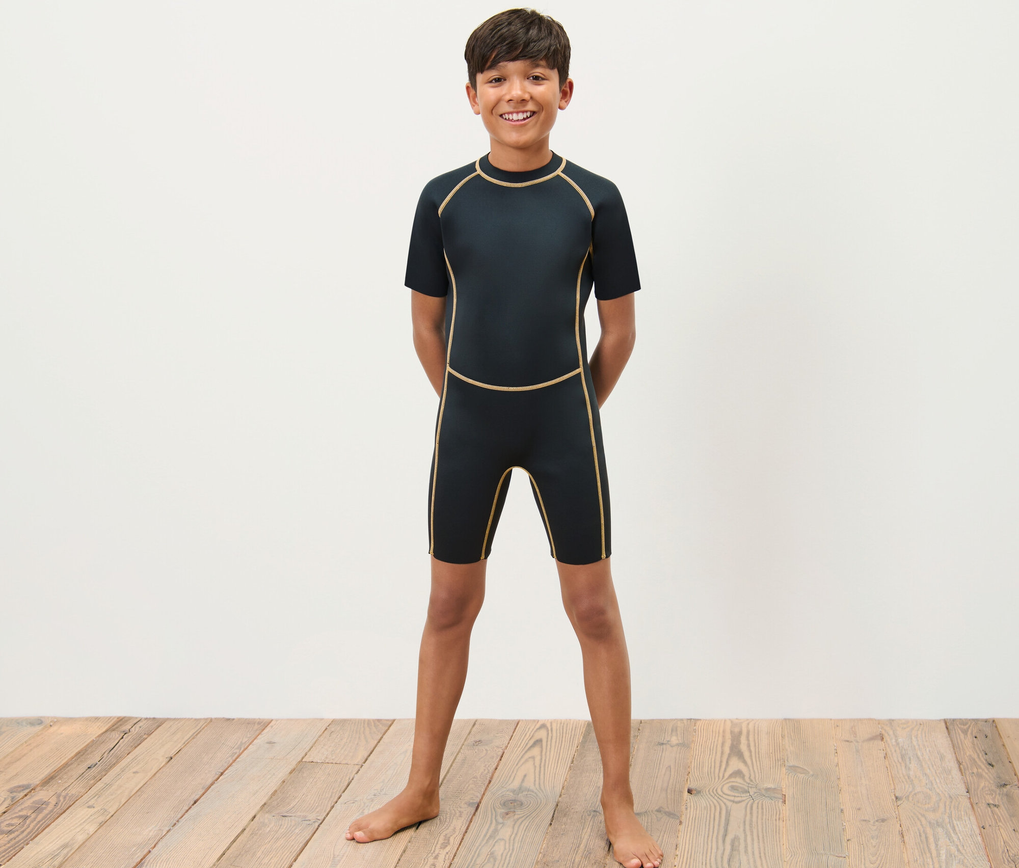 Ein Junge im Kinder-Neoprenanzug steht barfuß auf einem Holzboden vor einer weißen Wand.