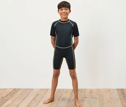 Ein Junge im Kinder-Neoprenanzug steht barfuß auf einem Holzboden vor einer weißen Wand.