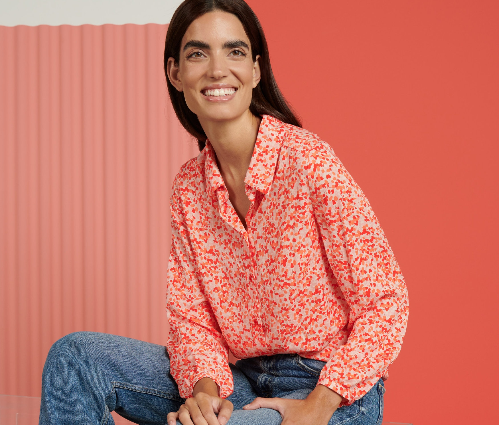 Eine Frau sitzt auf einem durchsichtigen Hocker und trägt eine Hemdbluse mit Print.