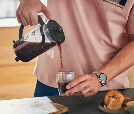 Mann gießt Kaffee aus einer schwarzen Siebstempelkanne 300 ml in ein Glas. Auf dem Tisch liegt ein Croissant.