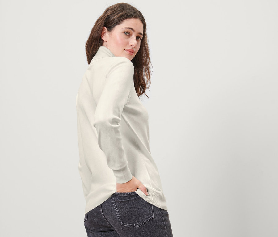 Frau posiert und blickt über ihre Schulter, sie trägt einen cremeweißen Feinstrick-Rollkragenpullover und Jeans.