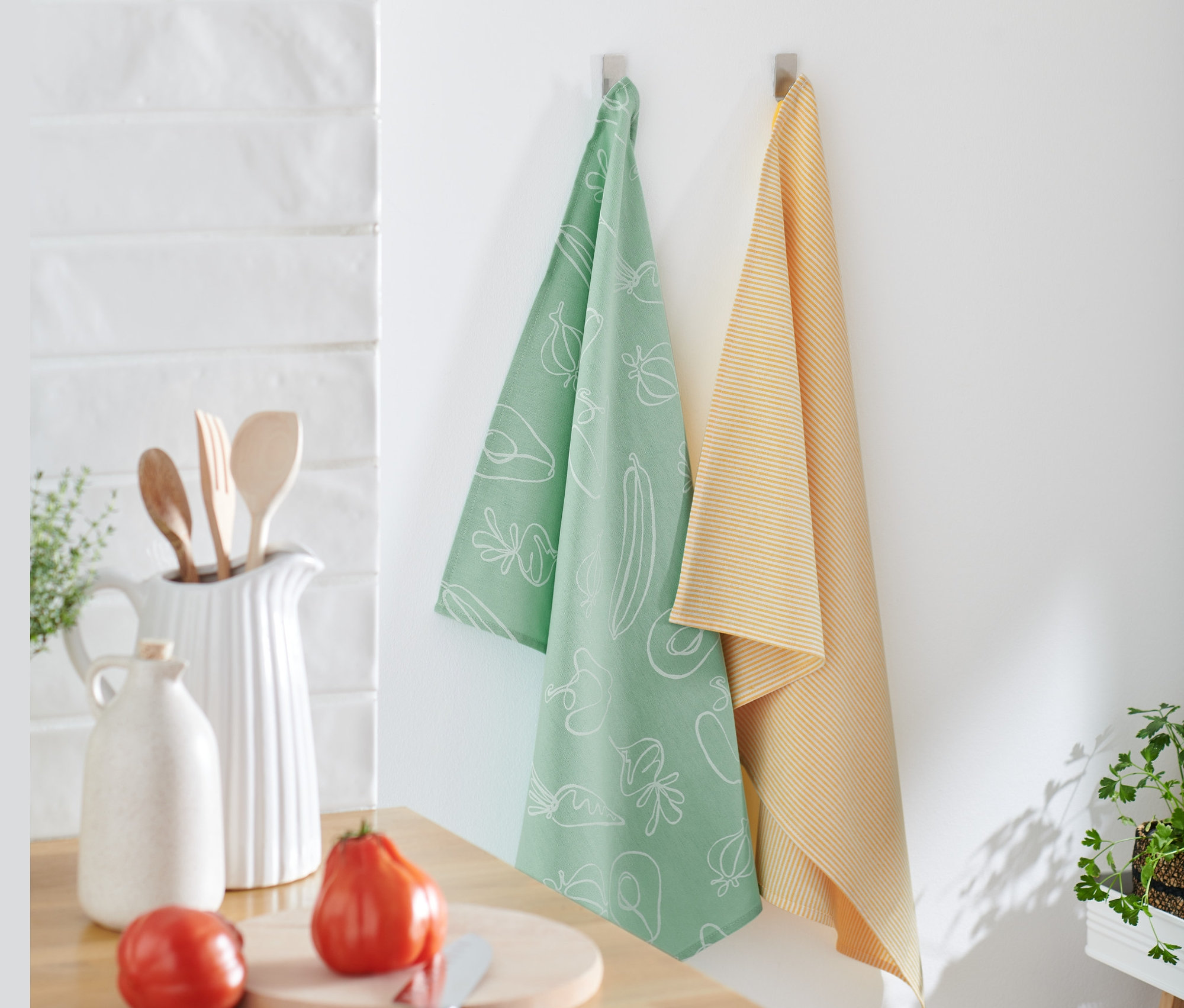 Zwei Geschirrtücher hängen an der Wand, eines hellgrün mit weißem Gemüseaufdruck und das andere gelb gestreift. 2 Geschirrtücher