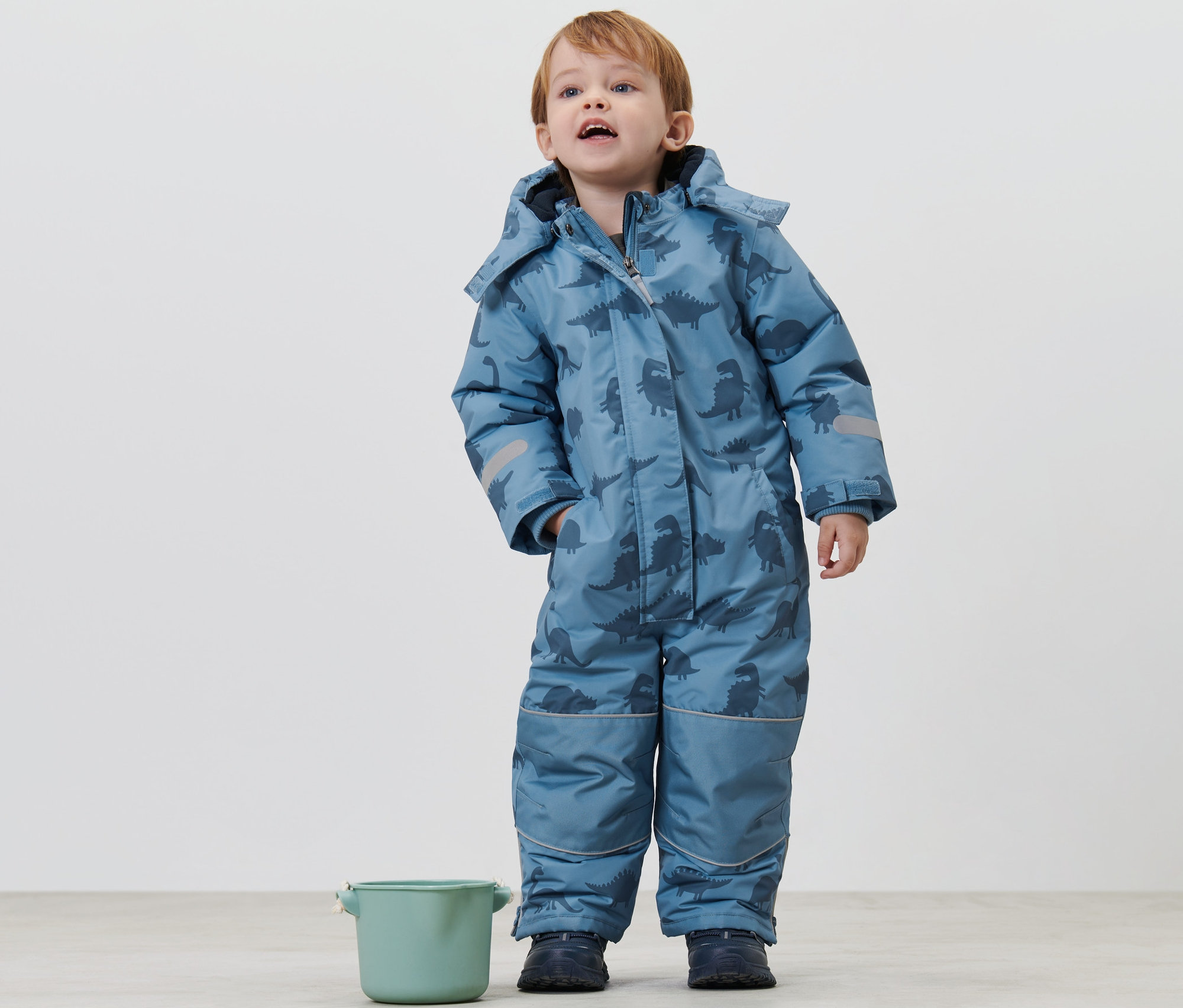Junge steht in Kinder-Schneeanzug mit Dinos und schaut nach oben. Neben ihm steht ein grüner Eimer.