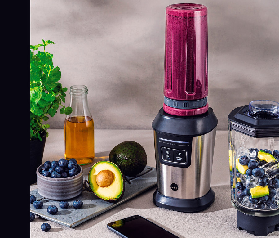 Auf einem Tisch stehen ein Wilfa Mixer »ACTIVLIFE B2GS-T« mit lila Smoothie, ein Glas mit Eis, Blaubeeren und Avocado. Auf dem Tisch liegen auch Avocado, Blaubeeren, Öl und ein Telefon.