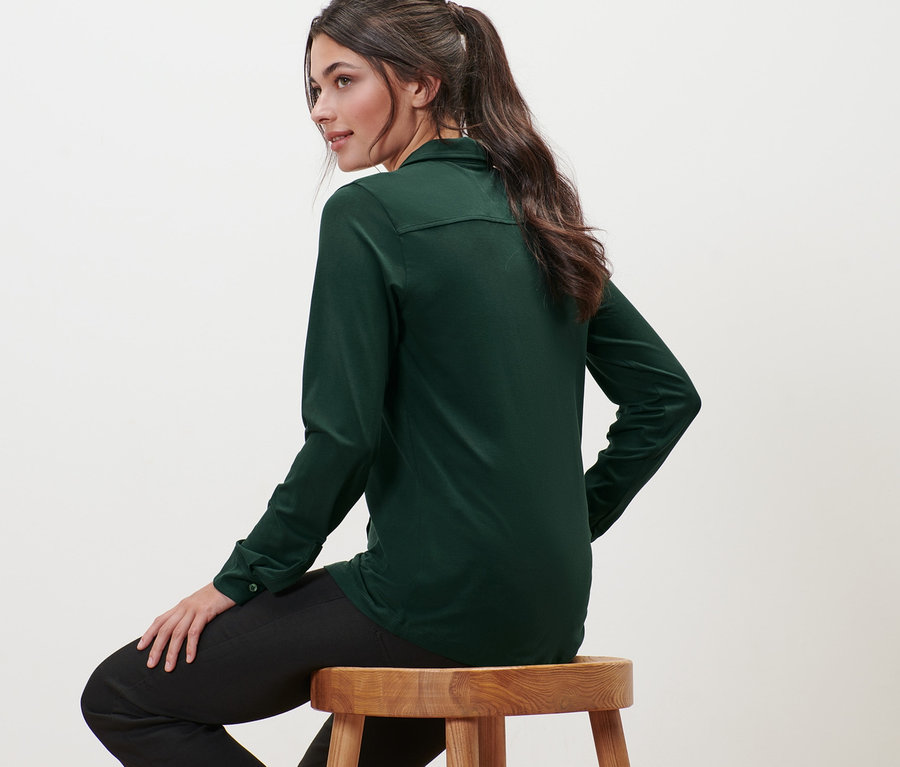 Eine Frau sitzt auf einem Holzhocker und trägt ein dunkelgrünes Blusenshirt mit Knopfleiste.