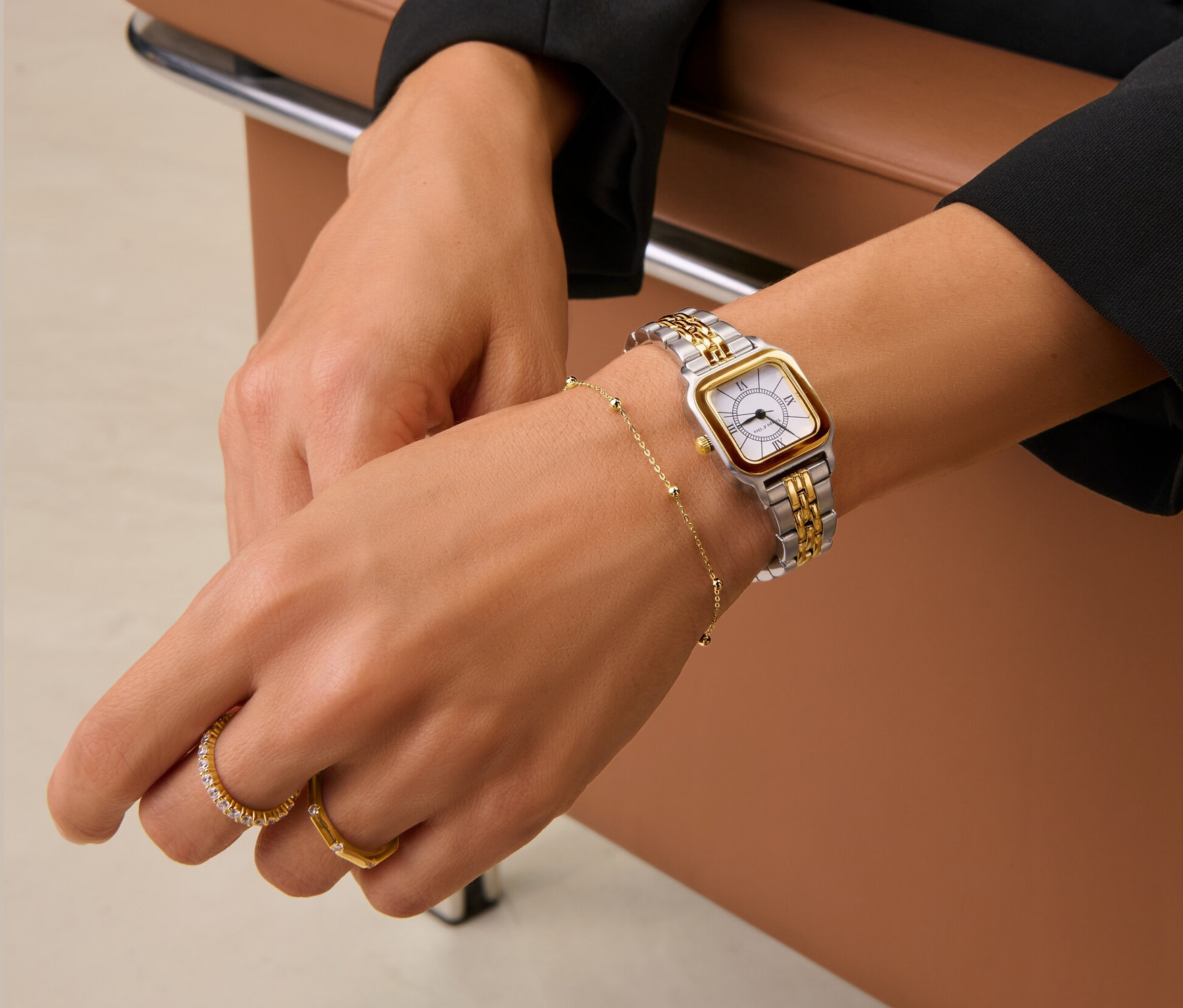 Eine Frau sitzt auf einem braunen Ledersessel und trägt eine Edelstahl-Bicolor-Damenuhr, ein goldenes Kugelarmband und zwei goldene Ringe, darunter einen 925 Silber Ring Memory vergoldet und einen 925 Silber Ring Octagon, vergoldet.