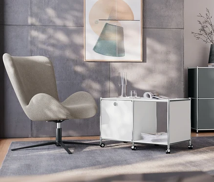 Im Wohnzimmer steht ein Drehsessel neben einem weißen Beistelltisch Metall »CN3« mit 1 Klappenfach und Rollen. Im Hintergrund befindet sich ein graues Sideboard Metall »CN3« mit 2 Klappenfächern und eine Tischleuchte aus Metall, verchromt.