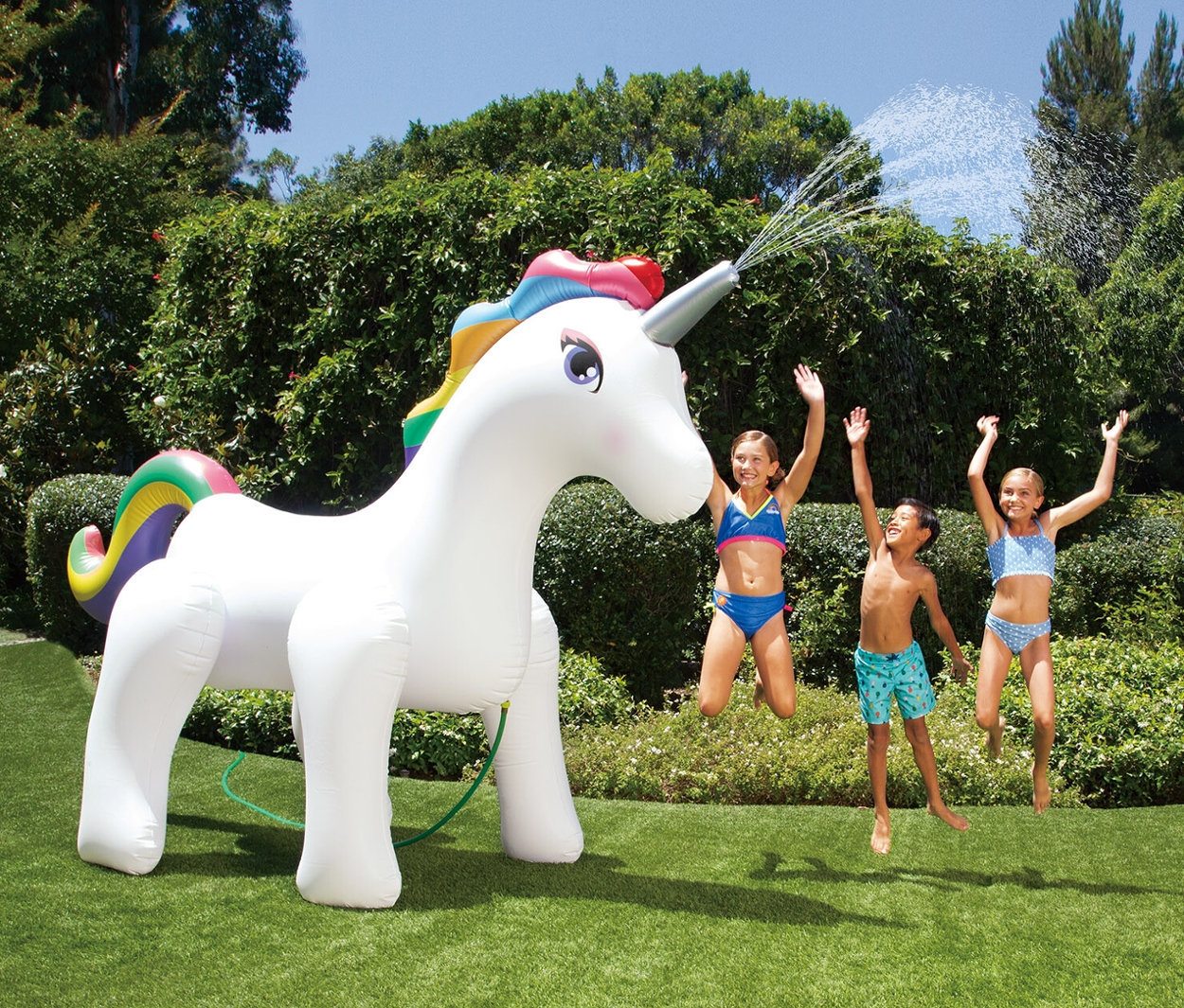 Drei Kinder springen auf einer Wiese neben einem großen aufblasbaren Summer-Waves-Sprinkler »Einhorn«, aus dem Wasser spritzt.