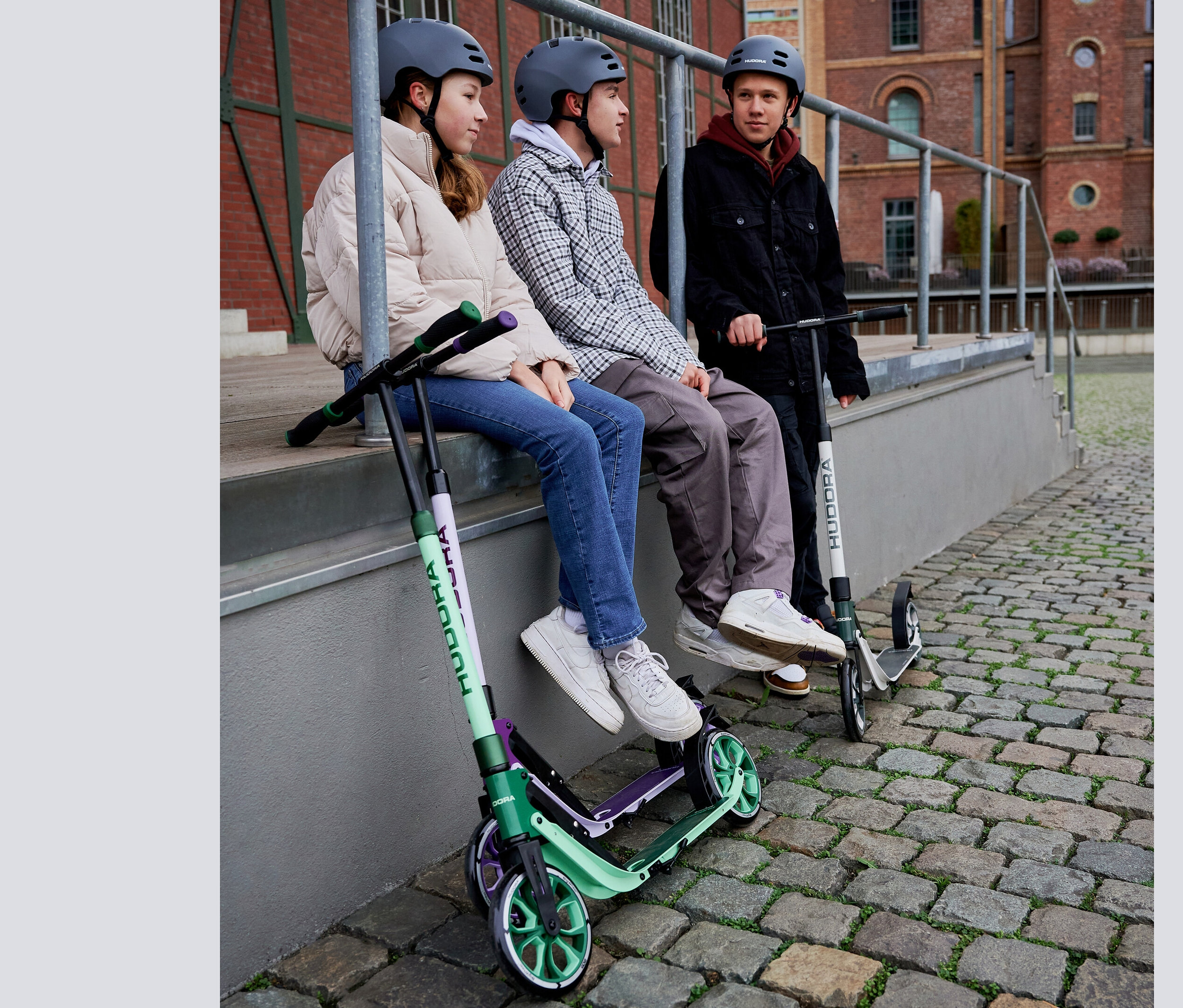 Drei Jugendliche sitzen nebeneinander auf einer Mauer. Neben ihnen stehen grüne HUDORA BigWheel® 205 Scooter.