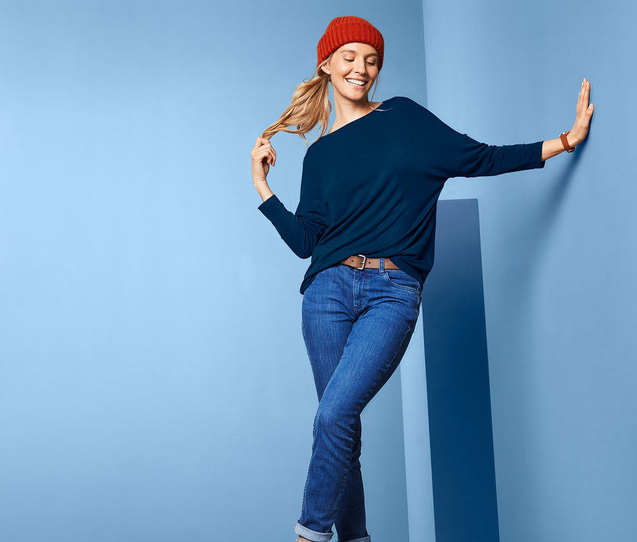 Frau posiert in blauem Oversized-Shirt, Jeans und roter Mütze vor blauem Hintergrund.