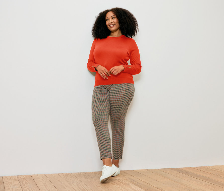 Frau posiert in orangefarbenem Feinstrickpullover, gemusterten Hosen und weißen Schuhen.