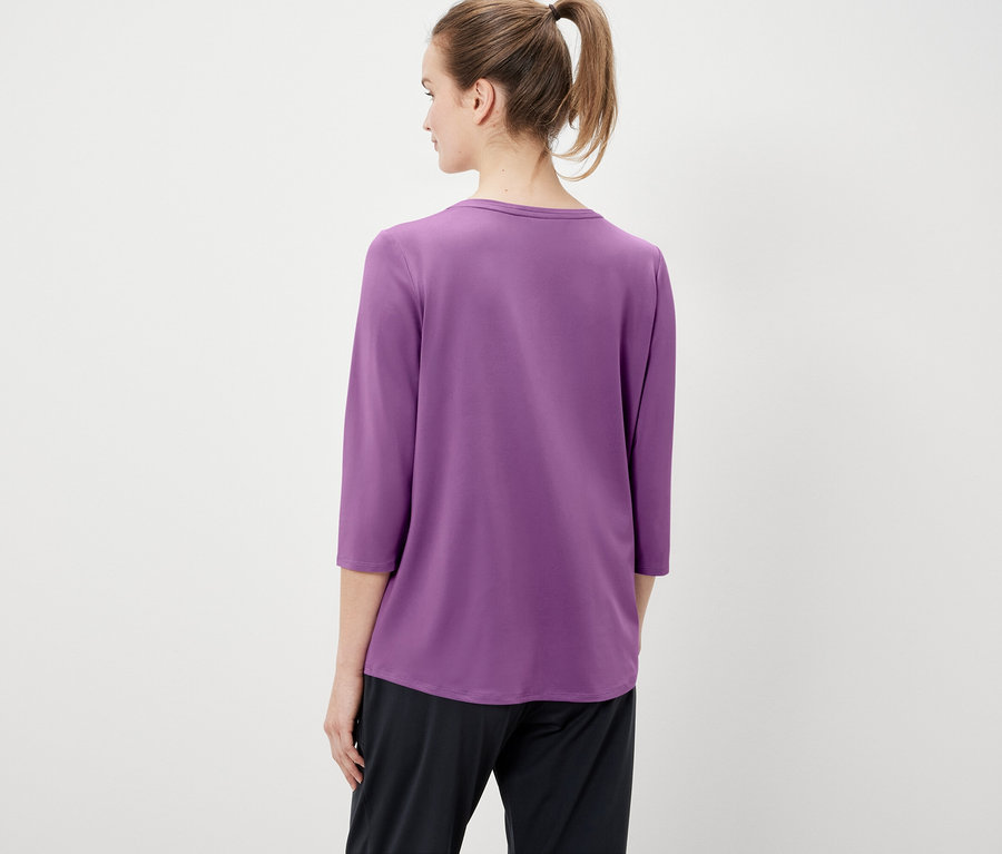 Von hinten ist eine Frau mit zum Pferdeschwanz gebundenen Haaren zu sehen, die ein violettes Dreiviertelarm-T-Shirt und eine schwarze Hose trägt.