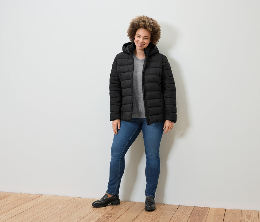 Eine Frau steht und lächelt in einer schwarzen Daunenjacke, blauen Jeans und schwarzen Loafern.