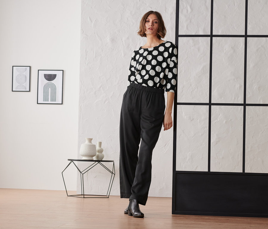 Frau posiert in schwarzer Hose und einem schwarz-weiß gepunkteten Shirt mit 3/4-Arm.