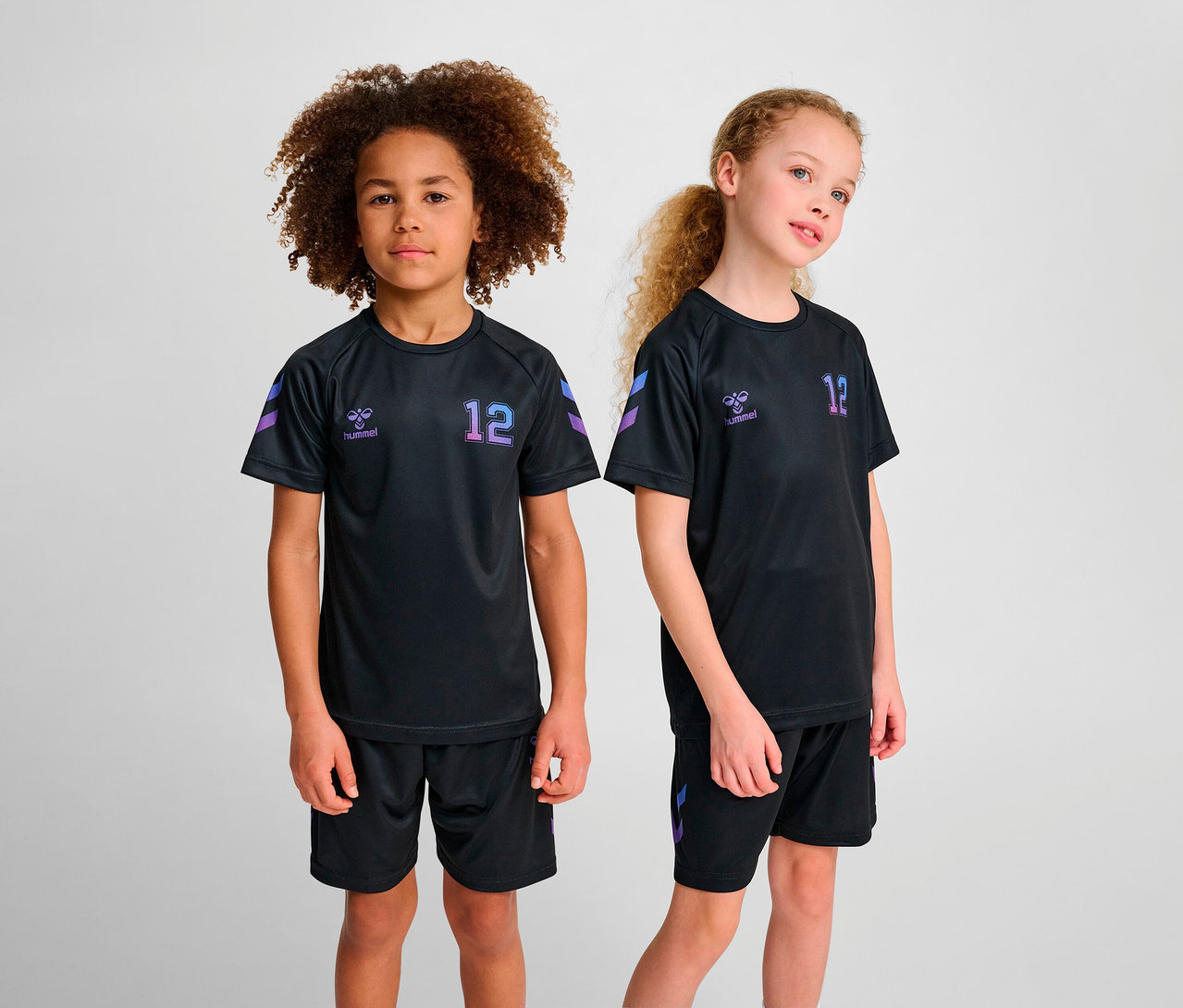 Zwei Kinder posieren in schwarzen Sporttrikots und Shorts. Beide tragen ein HUMMEL HMLJR shimmer T-Shirt S/S.