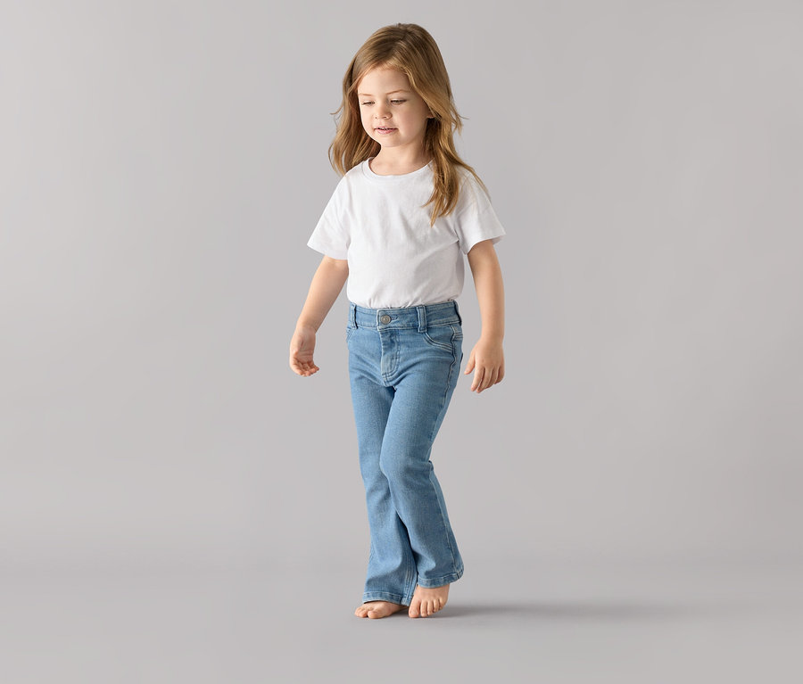 Ein Mädchen in einem weißen T-Shirt und der Kinder-Jeans ‒ Fit »Emma« steht barfuß da.