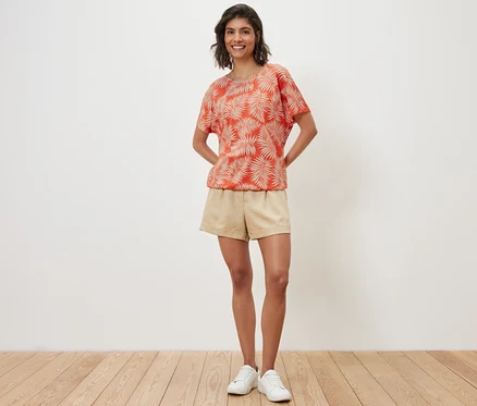 Eine Frau posiert in einem bedruckten Blusenshirt, Shorts und Turnschuhen.