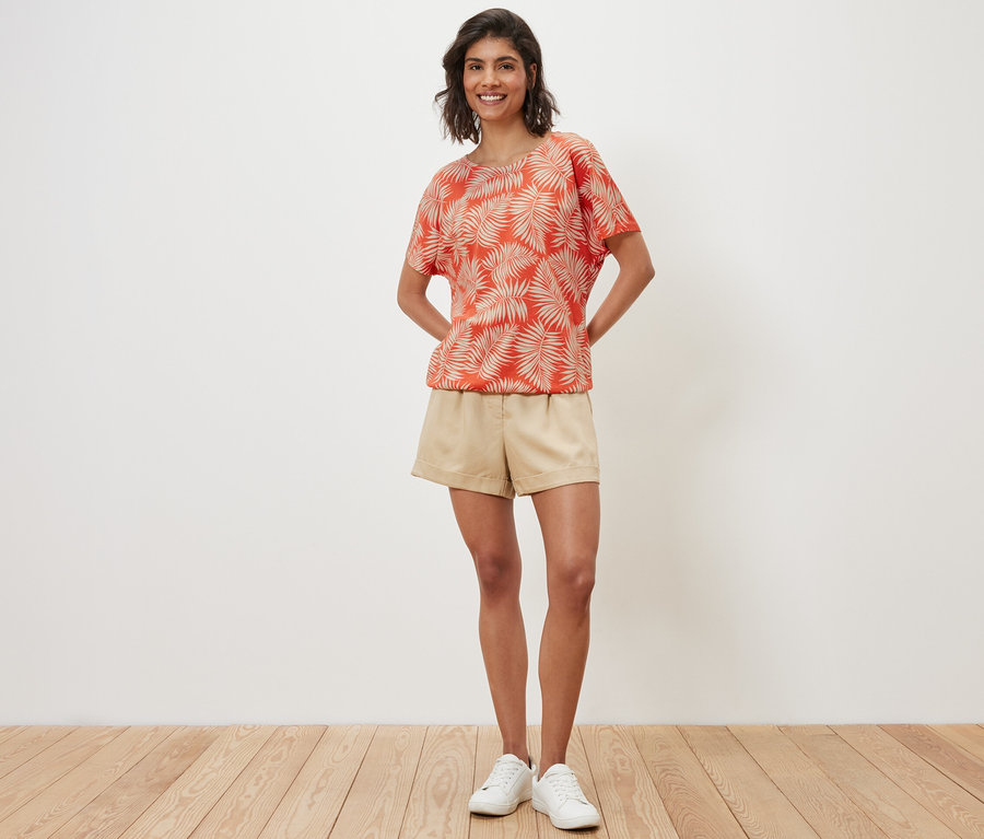 Eine Frau posiert in einem bedruckten Blusenshirt, Shorts und Turnschuhen.