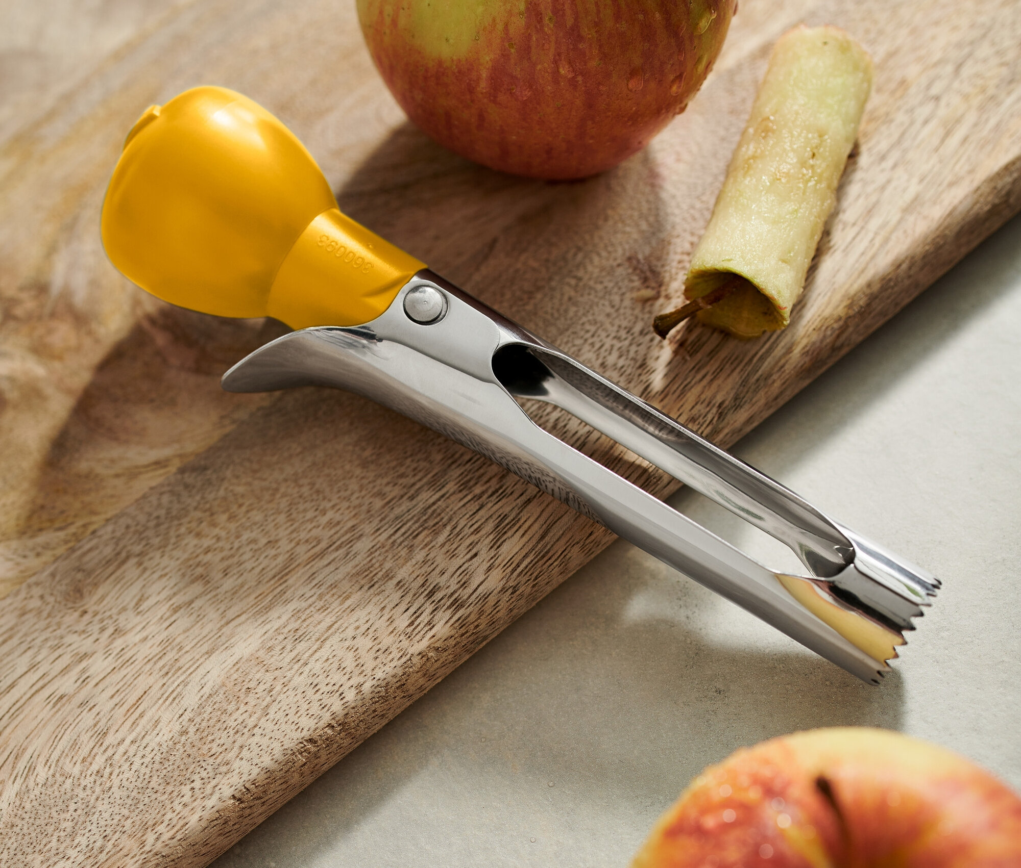 Auf einem Holzbrett liegen zwei Äpfel, einer mit entferntem Kern, daneben ein Stück Schale und ein Edelstahl-Kernentferner mit gelbem Griff.