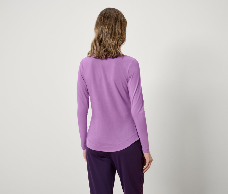 Rückansicht einer Frau mit gewelltem Haar in einem lavendelfarbenen Langarmshirt und lilafarbenen Hosen.