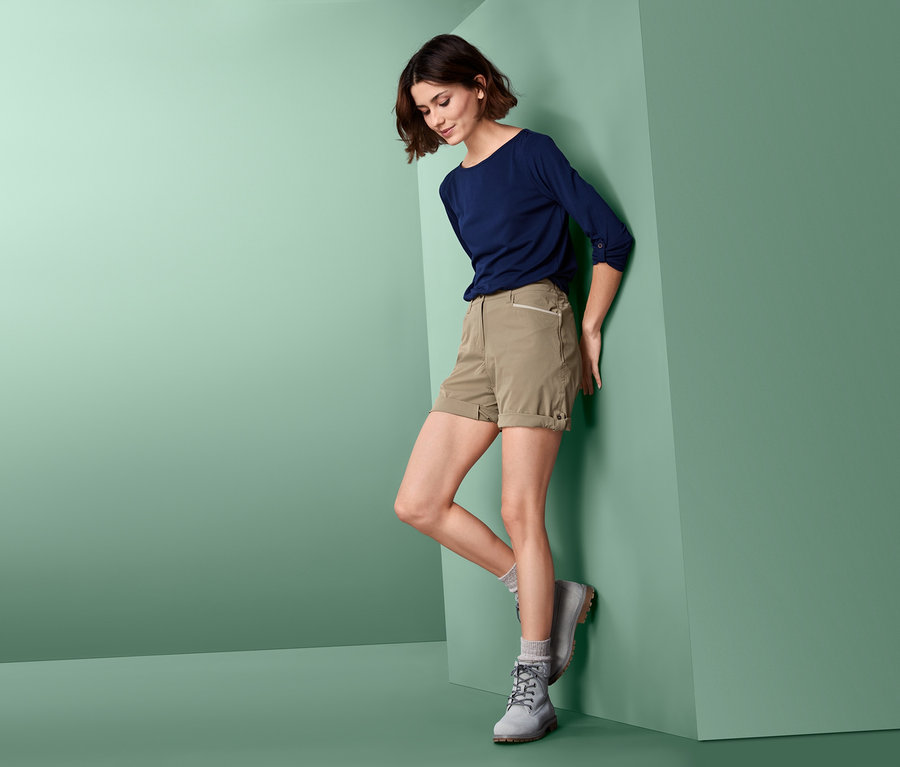 Eine Frau, die sich an eine Wand lehnt, trägt Softshell-Funktionsshorts, ein blaues T-Shirt und graue Stiefel.