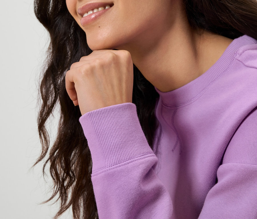 Nahaufnahme einer Frau mit gewelltem, dunklem Haar, die ihr Kinn mit der Hand abstützt und einen hellvioletten Pullover trägt.