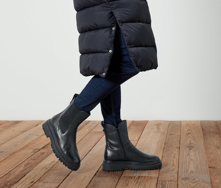 Beine einer Person in Jeans und schwarzen Lederstiefeln auf einem Holzboden. Die Person trägt eine lange, schwarze Jacke.