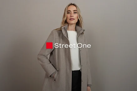 Frau mit blonden Haaren trägt einen beigen Mantel über einem weißen Oberteil und einer schwarzen Hose.