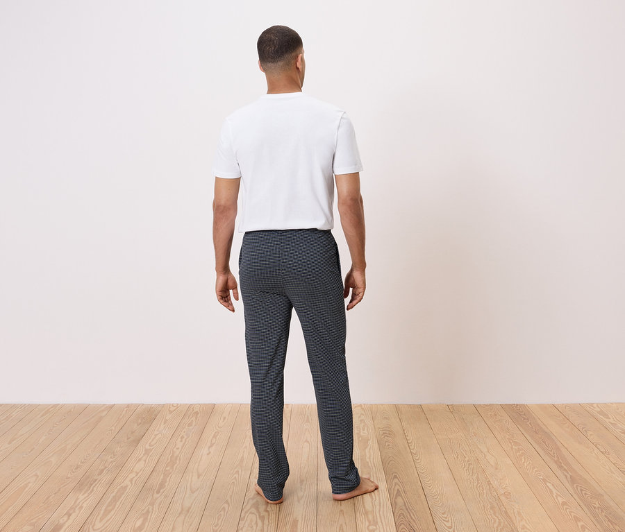 Ein Mann mit dem Rücken zur Kamera steht auf einem Holzboden und trägt ein weißes T-Shirt und eine dunkel karierte Hose.