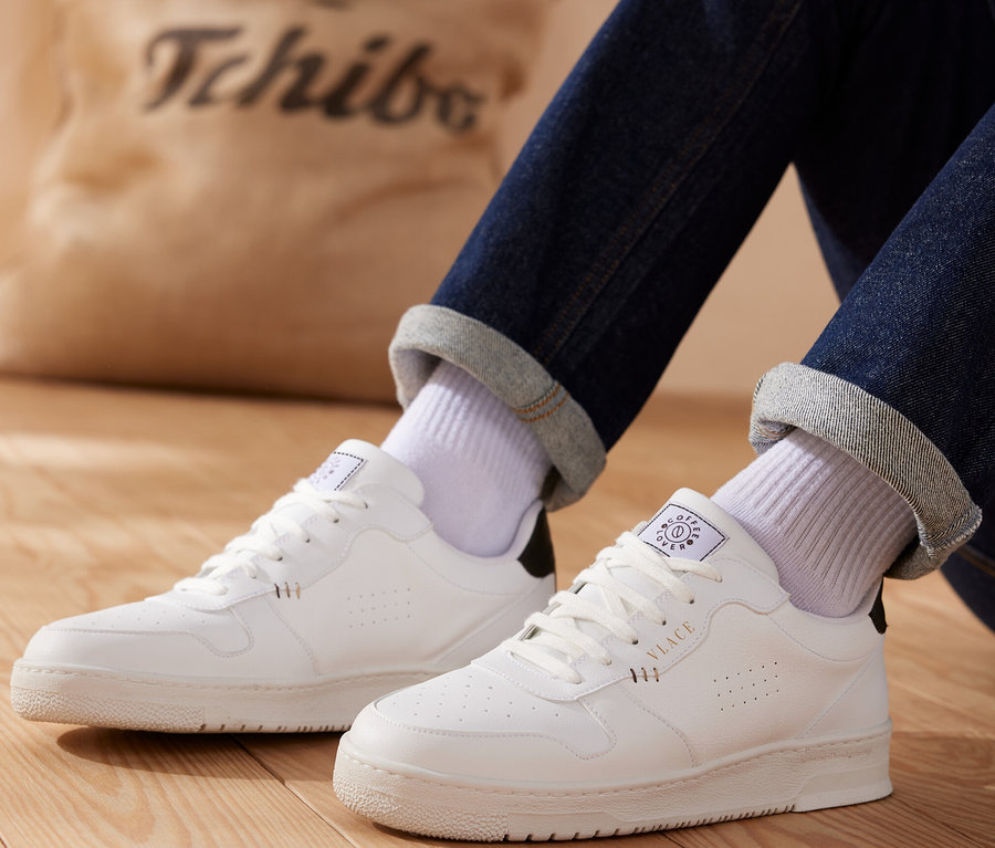 Beine in Jeans und weißen Socken, die weiße Tchibo x VLACE Coffee Lover Sneaker, Men, tragen, sitzen auf einem Holzboden. Im Hintergrund befindet sich eine beige Tasche mit dem Tchibo-Logo.