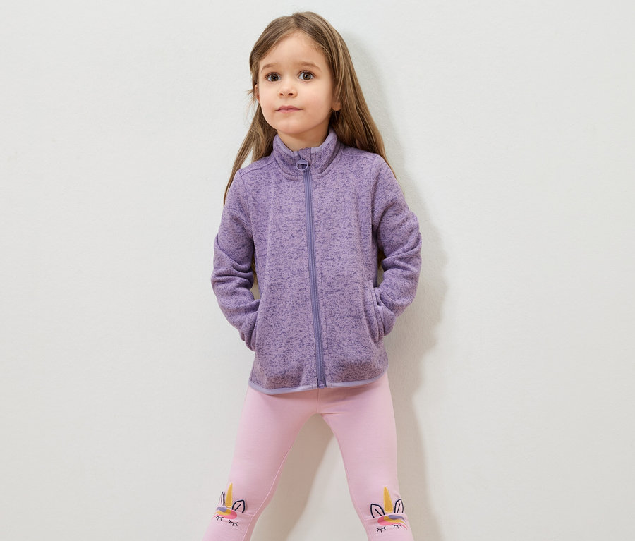 Mädchen mit braunen Haaren, lila Fleecejacke und rosa Leggings mit Einhornmotiv.