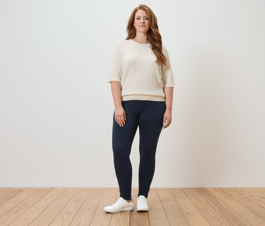Eine Frau steht in einem hellen Raum und trägt einen cremefarbenen Pullover, marineblaue Leggings und weiße Turnschuhe.
