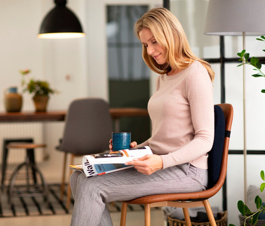 Frau sitzt auf einem Stuhl mit TEMPUR® Lordosekissen, liest eine Zeitschrift und hält eine Tasse.