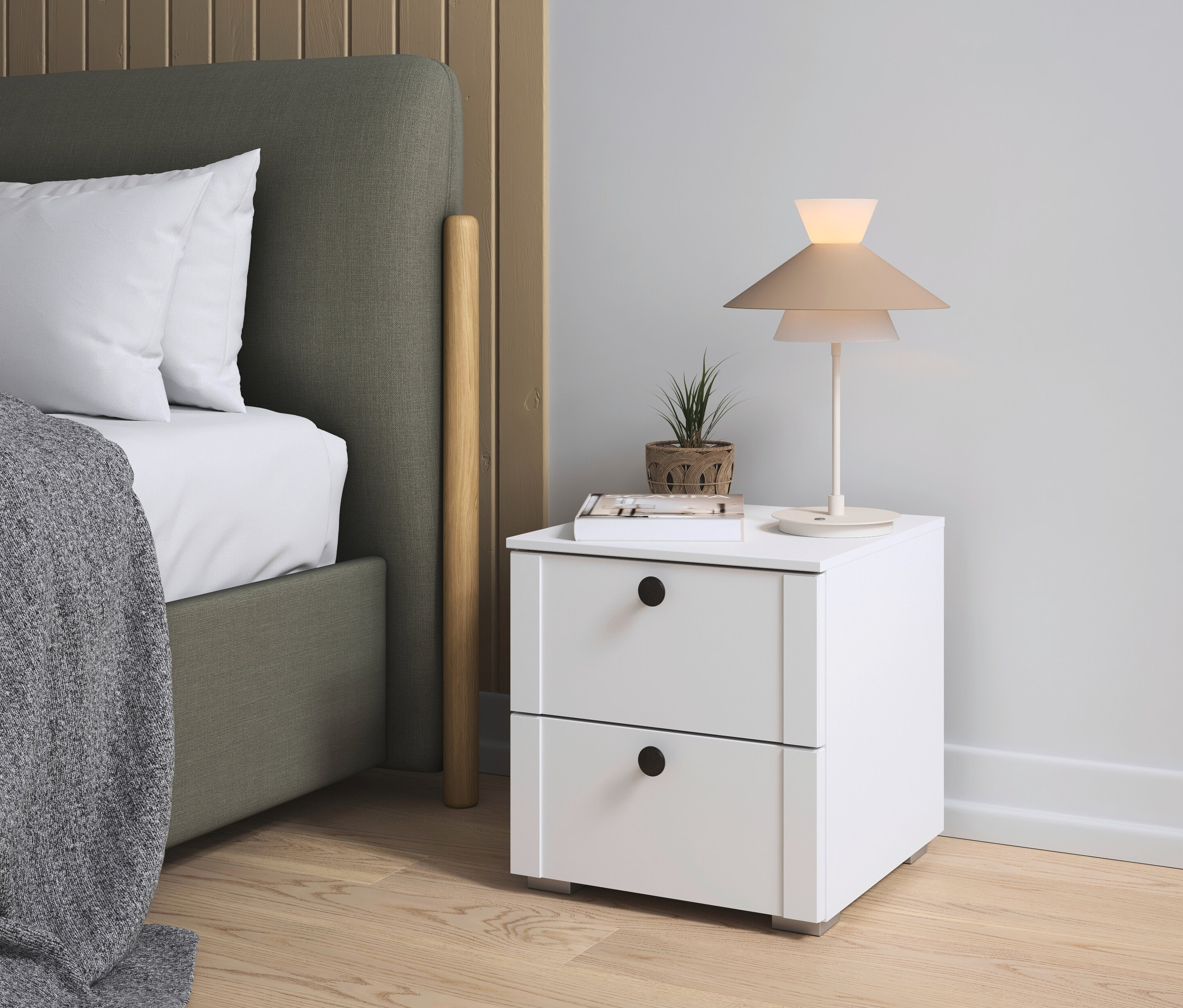 Nachttisch mit Lampe, Buch und Pflanze neben einem Bett mit grauer Tagesdecke und grünem Kopfteil.